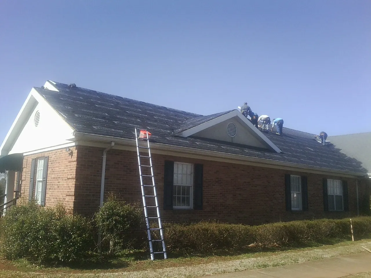 Crew tearing shingles off a brick ranch roof with a ladder set — Emergency Roof Repair work by our crew in Harvard